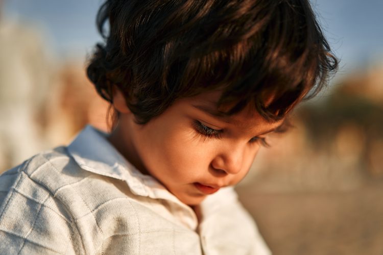 Little cute baby boy in white clothes playing with shells on a sandy beach by the sea.