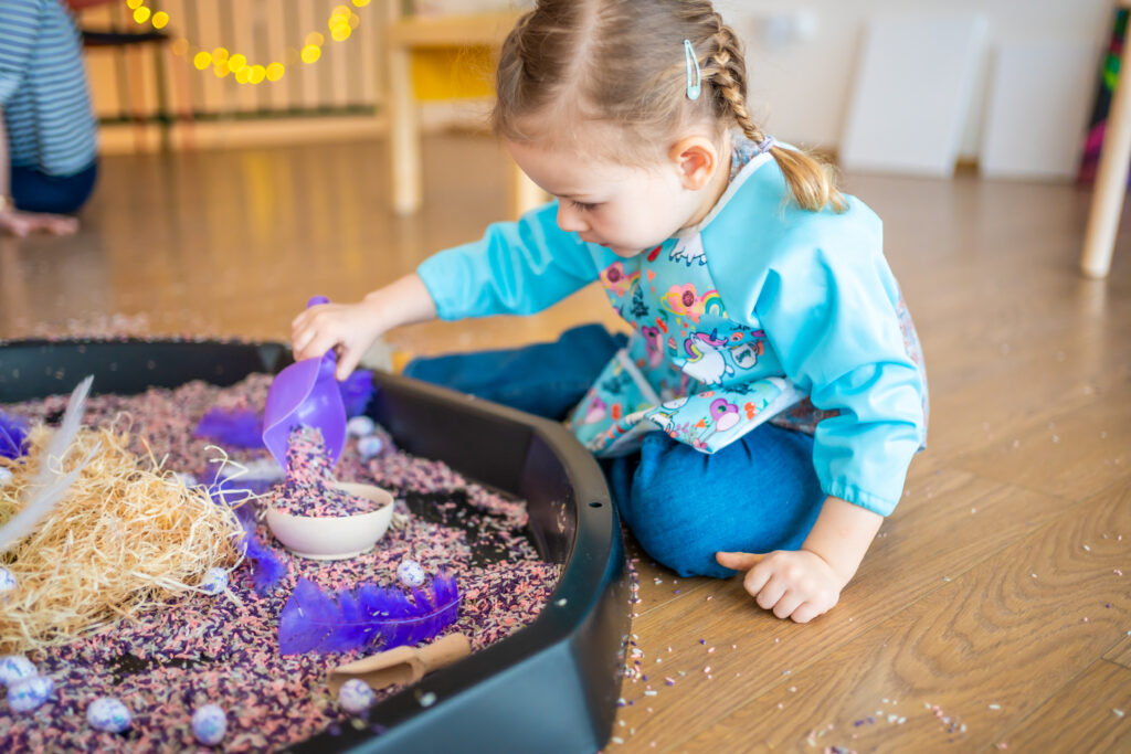 girl playing with a sensory bin filled with colorful rice, feathers, bowls and scoops